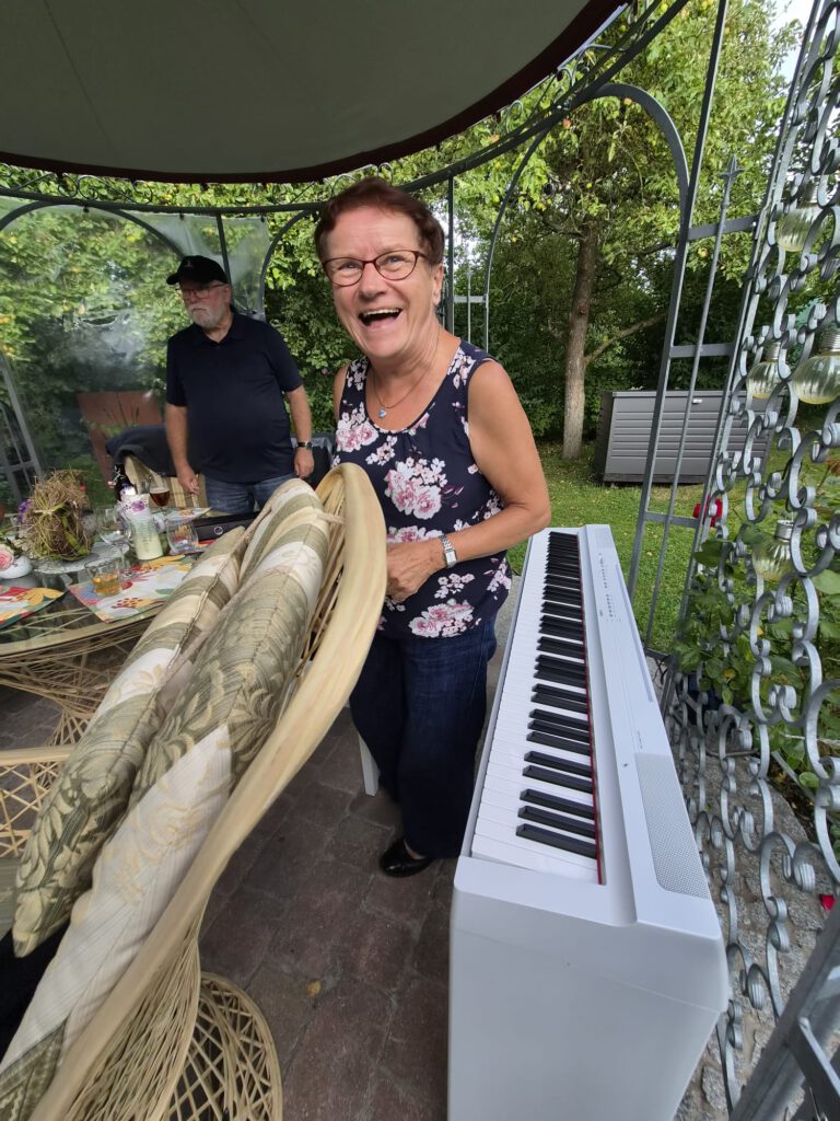 Christiane steht lachend hinter dem weißen Klavier im Gartenpavillon